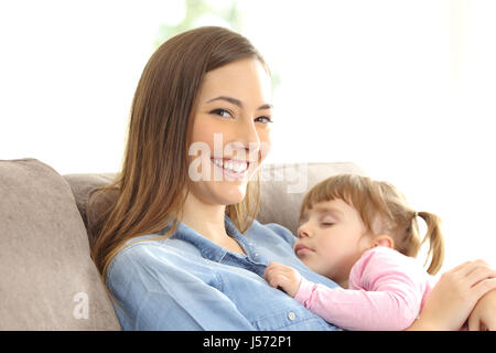 Mother looking at you with her daughter sleeping over her chest sitting on a couch at home Stock Photo