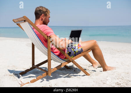 Full length of man using laptop while sitting on deck chair at beach Stock Photo
