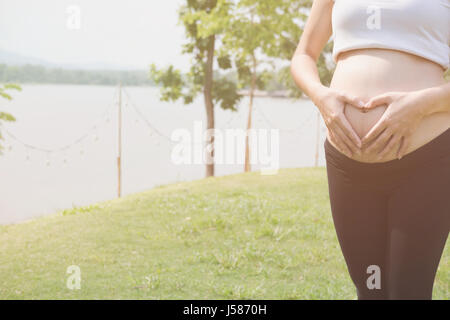 young pregnant woman making heart shape sign on her tummy in public park.  pregnancy, maternity belly care concept Stock Photo