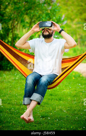 Young bearded man wearing virtual reality goggles relaxing in a garden hammock. Lifestyle VR fun and relax concept Stock Photo