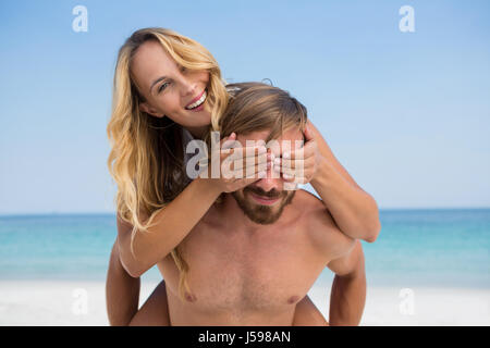 Man piggybacking playful woman while standing at beach against clear blue sky Stock Photo