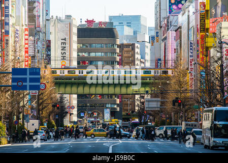 Looking down the wide street in Akihabara Electric City, Tokyo, Japan, during on busy daytime. People cross while a metro sails overhead Stock Photo