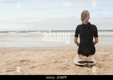 Rear view of woman kneeling on surfboard against cloudy sky Stock Photo