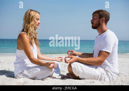 Side view of couple practicing meditation while sitting face to face at beach Stock Photo