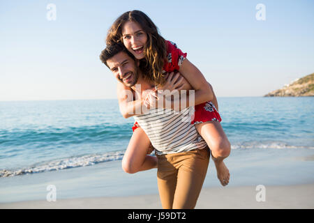 Portrait of young man piggybacking his cheerful girlfriend at beach on sunny day Stock Photo