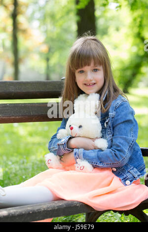 Little girl sits on a bench  in the park and holding in the hands of her favorite stuffed toy Stock Photo