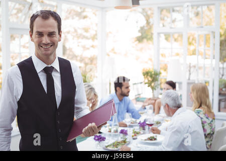 Portrait of smiling waiter holding menu while friends dining in background in restaurant Stock Photo