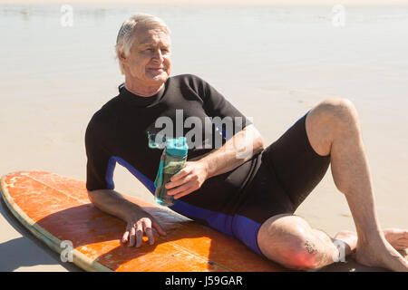 Senior man holding water bottle while sitting by surfboard at beach Stock Photo