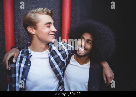 Cheerful male friends with arm around standing against wall Stock Photo