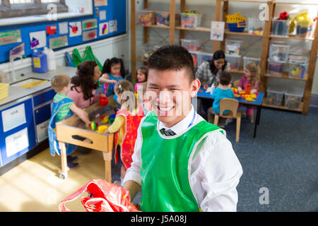 Teacher in a classroom. He is holding some aprons and smiling at the camera. The children are playing in the background with other teachers. Stock Photo