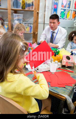 Little girl using scissors to cut shapes from a red piece of paper. She is in a nursery class with other students and a teacher. Stock Photo