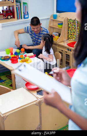 Male teacher playing in a toy kitchen with a nursery student. There is a teacher in the foreground with a clipboard who is watching them. Stock Photo