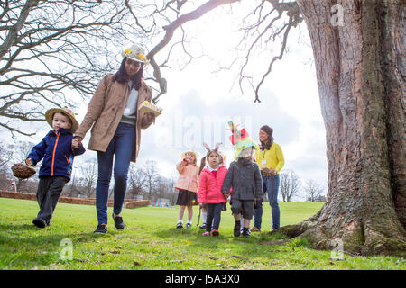 Nursery teachers and students doing an Easter egg hunt outdoors. They are wearing handmade hats and carrying baskets. Stock Photo