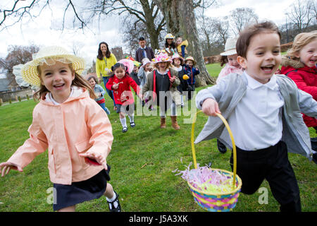 Nursery children running across a field during their outdoor Easter egg hunt, they are wearing handmade hats and carrying baskets. Stock Photo