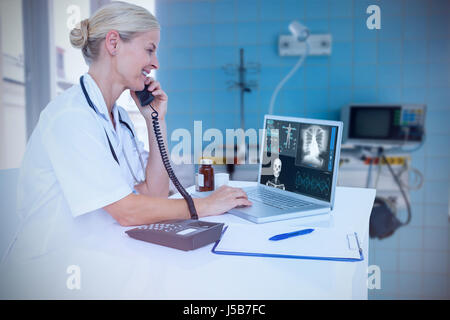Smiling doctor using laptop while talking on telephone against human skeleton with various medical xray reports Stock Photo