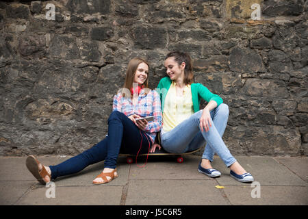 Two fashionable women sitting on skateboard and using a Smart Phone device. Stock Photo