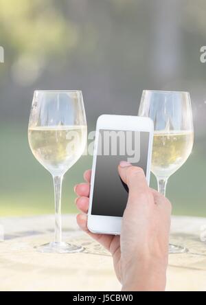 Digital composite of Hand holding phone with two glasses of champagne and sunny greenery Stock Photo