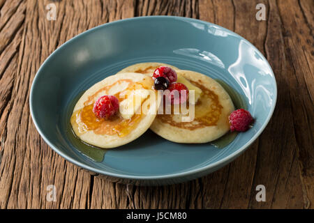 Three fresh pancakes, drizzled with syrup and Berries in a blue bowl, sitting on a rustic wooden table. Stock Photo