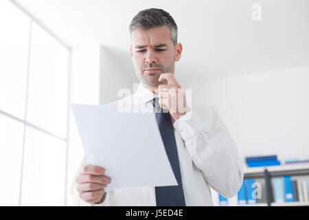 Businessman examining expensive bills on a document, he is frowning and touching his chin Stock Photo