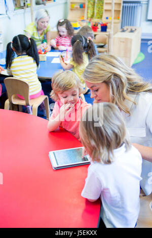 Female teacher using a digital tablet with her nursery students in a classroom. There is a small group of students working with another teacher in the Stock Photo