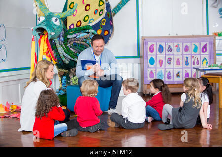 Small group of nursery children are sitting on the floor in the school hall around their teacher. He is holding books and talking to them. Stock Photo