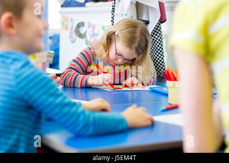 Little girl sitting at a table in her nursery classroom. She is colouring in with felt tip pens. Stock Photo