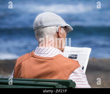 Elderly man wearing hearing aid reading sitting on bench overlooking the sea. Stock Photo