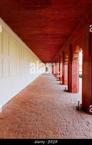Looking down a long red and white corridor on the outside of a building with open archways down left side and a wooden door in the far distance. Stock Photo