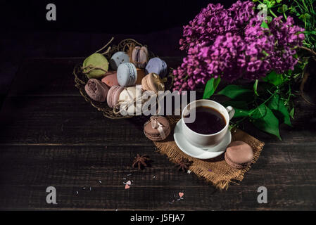 Cup of coffee on linen napkin and several multicolored macarons in braid old vase with branch of lilac. Stock Photo