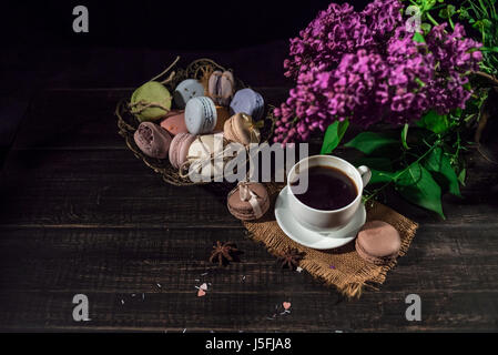 Cup of coffee on linen napkin and several multicolored macarons in braid old vase with branch of lilac. Stock Photo