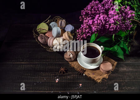 Cup of coffee on linen napkin and several multicolored macarons in braid old vase with branch of lilac. Stock Photo