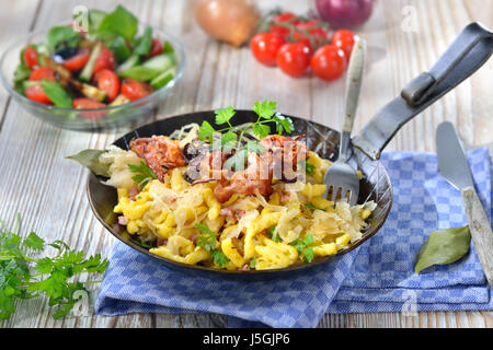 Swabian spaetzle with sauerkraut, fried bacon and onion rings on a wooden table with a side salad Stock Photo