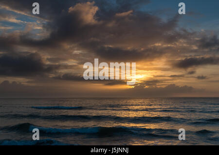 Sun hiding behind clouds during a beach sunrise with storm clouds rolling in Stock Photo