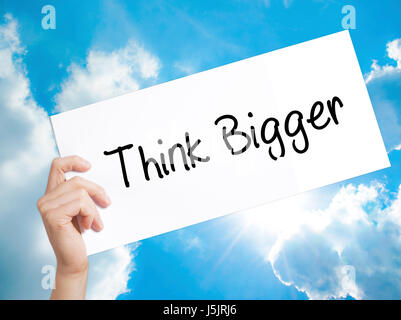 Think Bigger Sign on white paper. Man Hand Holding Paper with text. Isolated on sky background. Isolated on background. Business, technology, internet Stock Photo