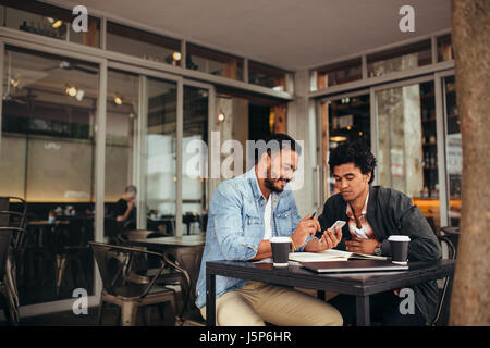 Two young men sitting together at coffee shop using mobile phone. Friends meeting at cafe and using smart phone. Stock Photo