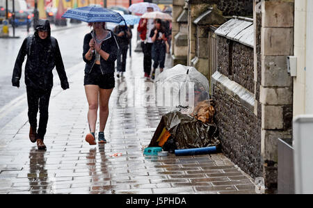 Brighton, UK. 18th May, 2017. A man asks for money under an umbrella on the pavement as people walk past in torrential rain in central Brighton today Credit: Simon Dack/Alamy Live News Stock Photo