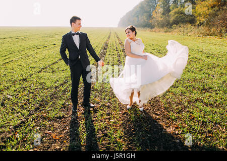 Lovely view of the newlyweds in the green field. The bride is playing with her wedding dress. Stock Photo