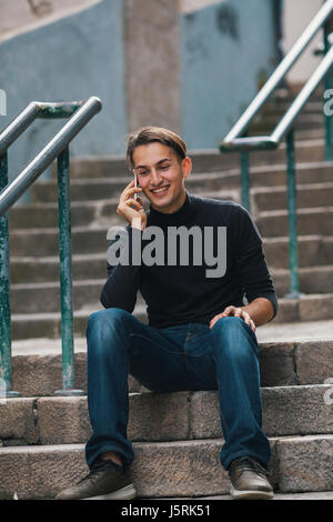 Handsome young man emotionally talking on the phone sitting on stone steps in old european downtown. Stock Photo