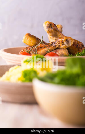 Vertical photo with baked or fried chicken legs on clay plate. Food is on wooden boarrd with grey textured background. Green salat and meshed potatoes Stock Photo