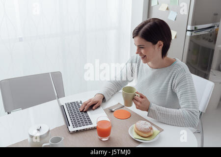 Young smiling woman having breakfast in the kitchen and social networking with a laptop Stock Photo