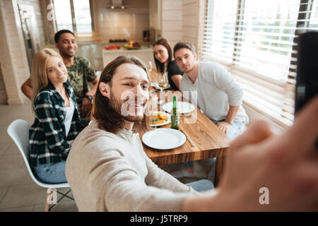 Attractive young man taking a selfie photo with friends in the kitchen while having lunch together at home Stock Photo