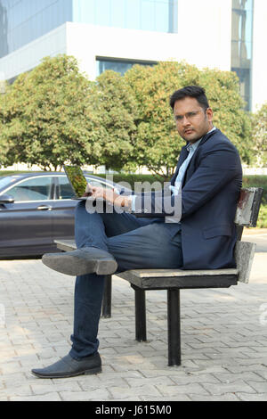 Young Indian corporate man in suit sitting on bench and working on his laptop Stock Photo