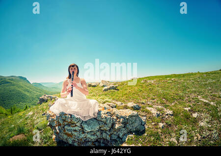 Young woman in white dress playing on vintage flute on the  a mountain landscape. Travelling concept art photo. Stock Photo