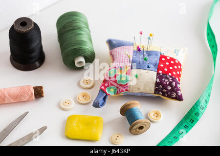 closeup sewing tools , patchwork, tailoring and fashion concept - thread spools, buttons, measuring tape, pincushion, scissors, pieces of colored patchwork fabric, soap on white desk. Stock Photo