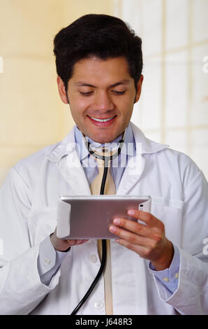 Handsome smiling doctor with an Stethoscope holding from his neck, checking something in his tablet, in office background Stock Photo