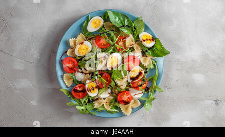 Farfalle bow tie pasta salad with quail eggs, cherry tomatoes, rucola, mozzarella and basil, dressed with balsamic sauce over grey concrete background Stock Photo