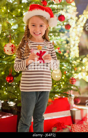 Portrait smiling girl in Santa hat holding gift in front of Christmas tree Stock Photo