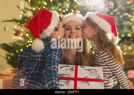 Daughter and son in Santa hats kissing mother with Christmas gift Stock Photo