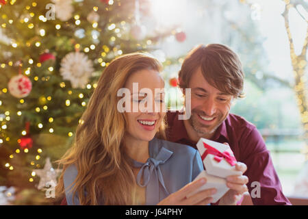 Husband watching wife opening Christmas gift in front of Christmas tree Stock Photo