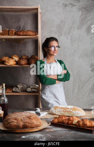 Picture of concentrated young lady baker standing with arms crossed at bakery near bread. Looking aside. Stock Photo